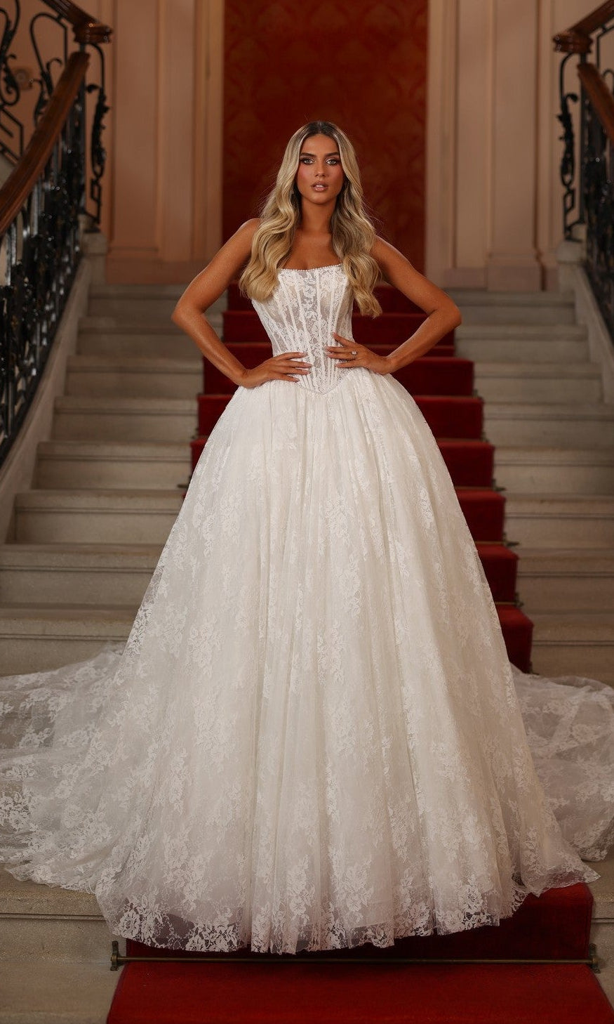 A woman in an ivory solid Long White Dress: Alyce 7149 stands on a grand red-carpeted staircase, hands on her hips, showcasing the elegant ball gown style.